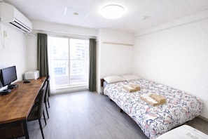 A hotel room with a wooden desk, a bed with floral bedding, a window with curtains, and an air conditioning unit.