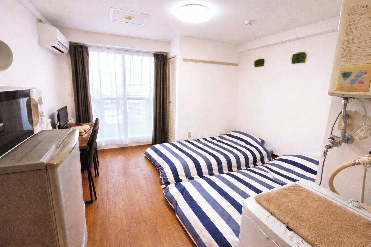 Une chambre d'hôtel avec une kitchenette, un lit avec une literie à rayures, un bureau avec une chaise et une fenêtre avec des rideaux.