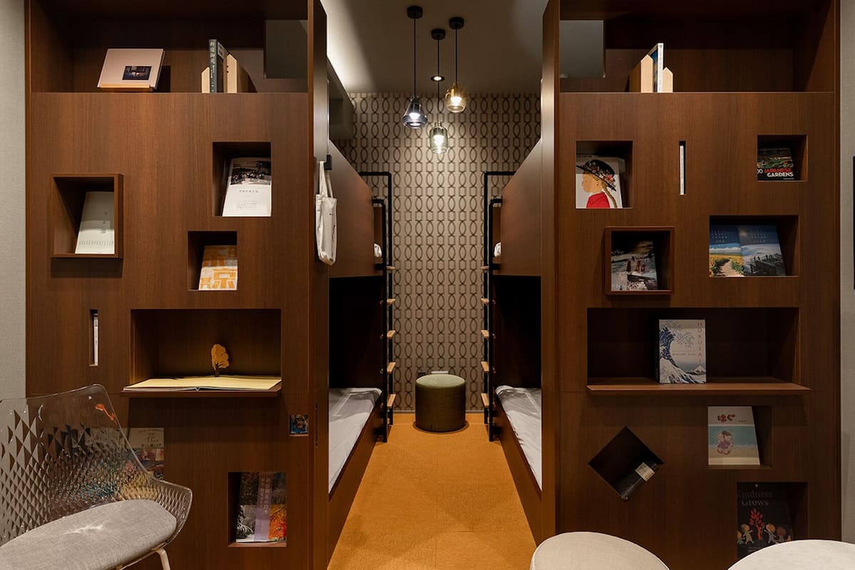 A hotel corridor with wooden shelving units displaying books and magazines, a central round table, and a seating area with chairs.