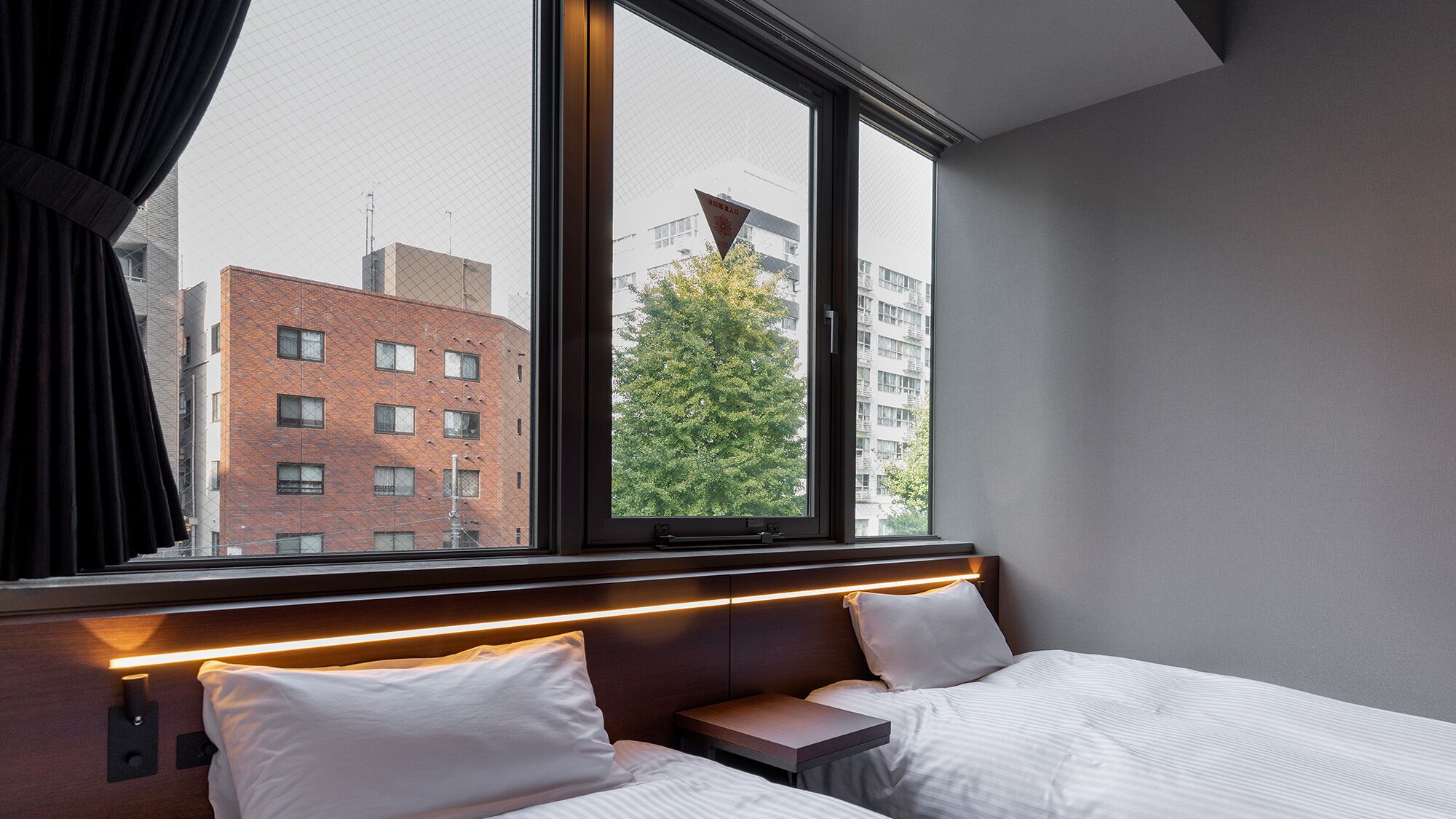 A hotel room with two beds, a window with a view of a brick building and a tree, and a headboard with built-in lighting.