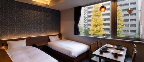 A hotel room with two beds, a dining table with chairs, and a view of a cityscape through the window.