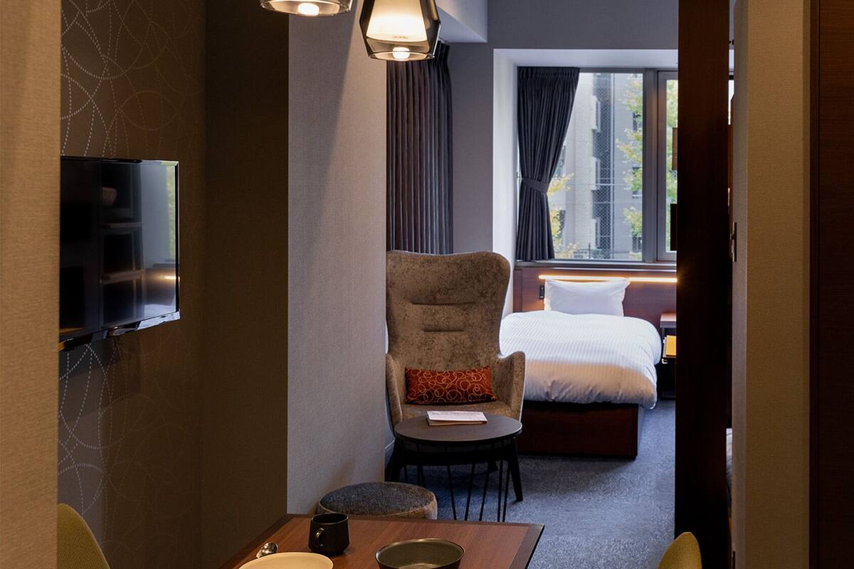 Een hotelkamer met een aan de muur bevestigde televisie, een stoel met kussen, een klein tafeltje met mok, en een bed met hoofdeinde.