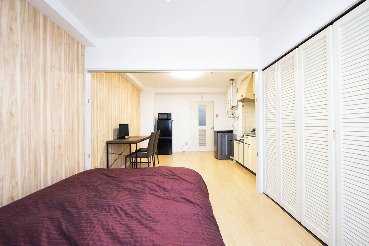 A compact room with a bed, a small dining table with chairs, a kitchenette, and a wardrobe.