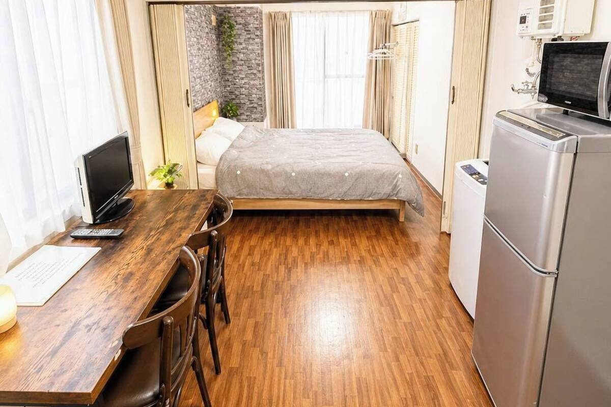 A compact hotel room with a wooden floor, a bed with a grey bedspread, a desk with a laptop, a small refrigerator, and a window with curtains.