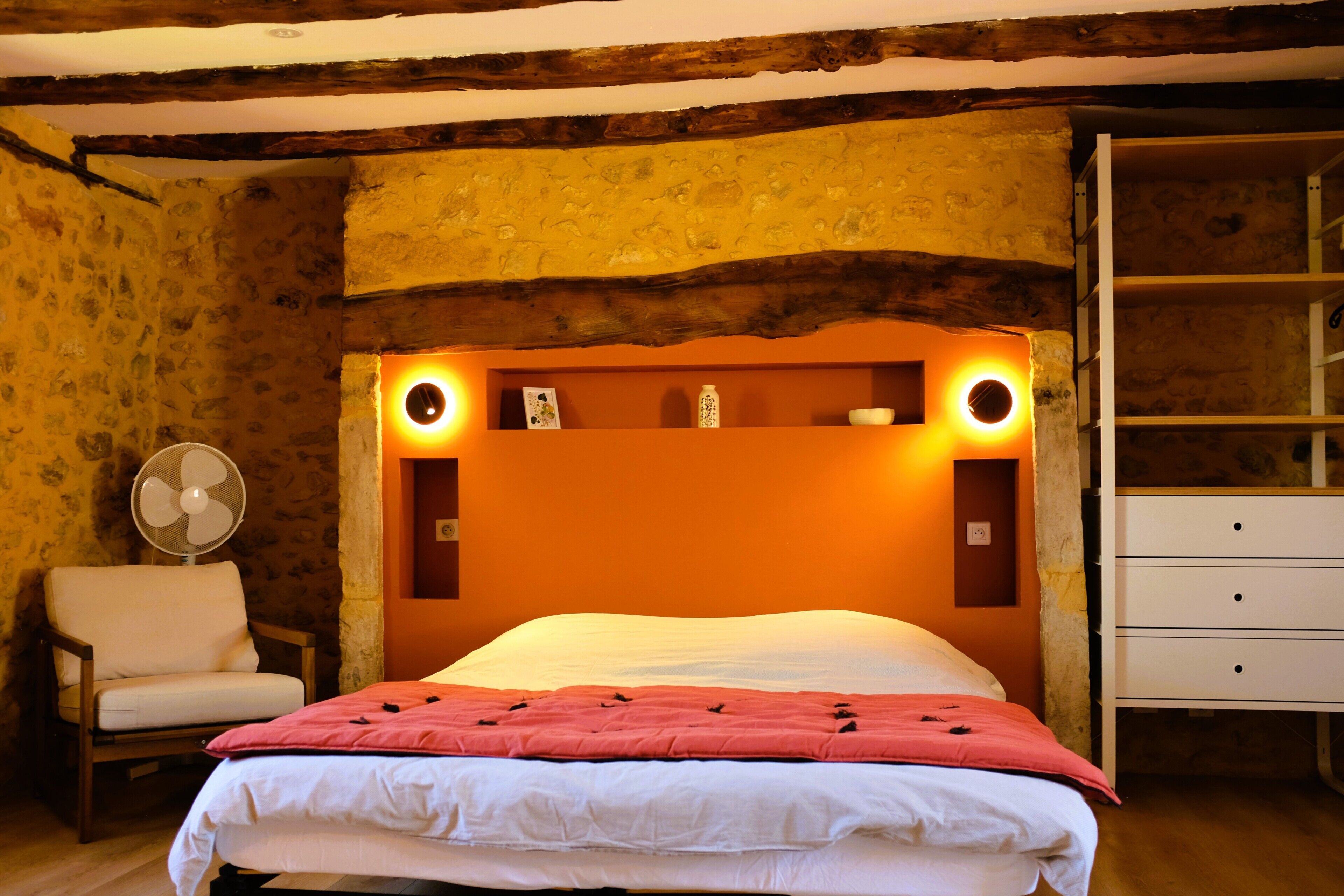 A bedroom with a wooden headboard, a bed with white bedding, a chair, a bedside table, and a fan.