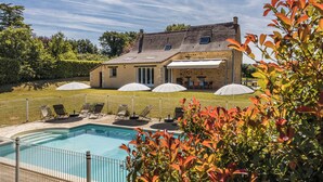 Une piscine entourée de chaises longues et de parasols, adossée à une maison en pierre avec un toit en tuiles et un jardin clos.