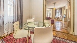 Un salon moderne avec une table basse au plateau en verre, des chaises blanches, un tapis à motifs rouges et une vue sur la salle à manger grâce à une porte ouverte.