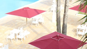 A poolside area with white chairs and red umbrellas, surrounded by palm trees and a tiled floor.