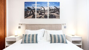 A neatly made bed with white linens and striped pillows, flanked by two bedside tables with lamps, and a large beach-themed artwork above.
