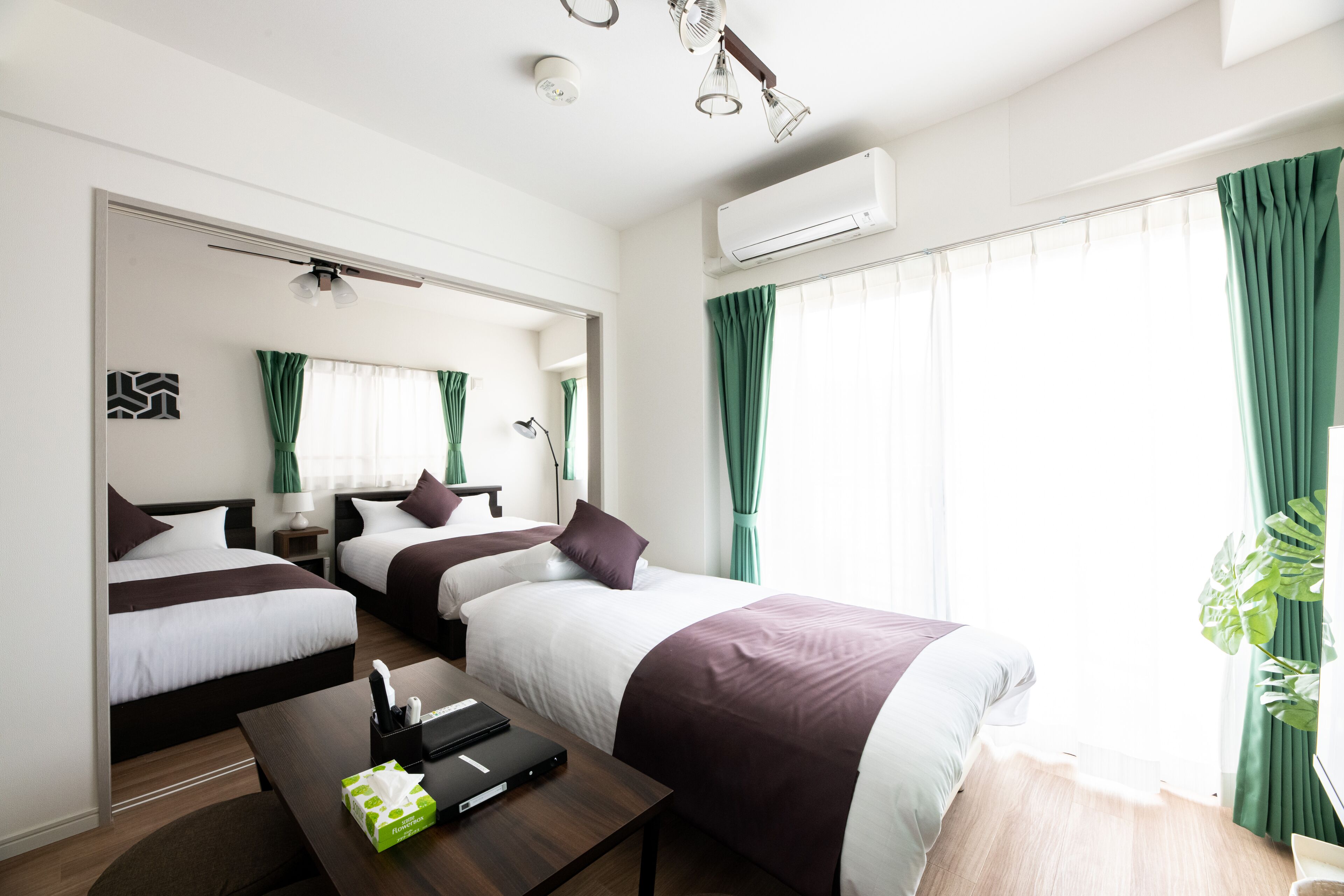 A hotel room with two beds, a desk, a mirror, an air conditioner, and a window with curtains.
