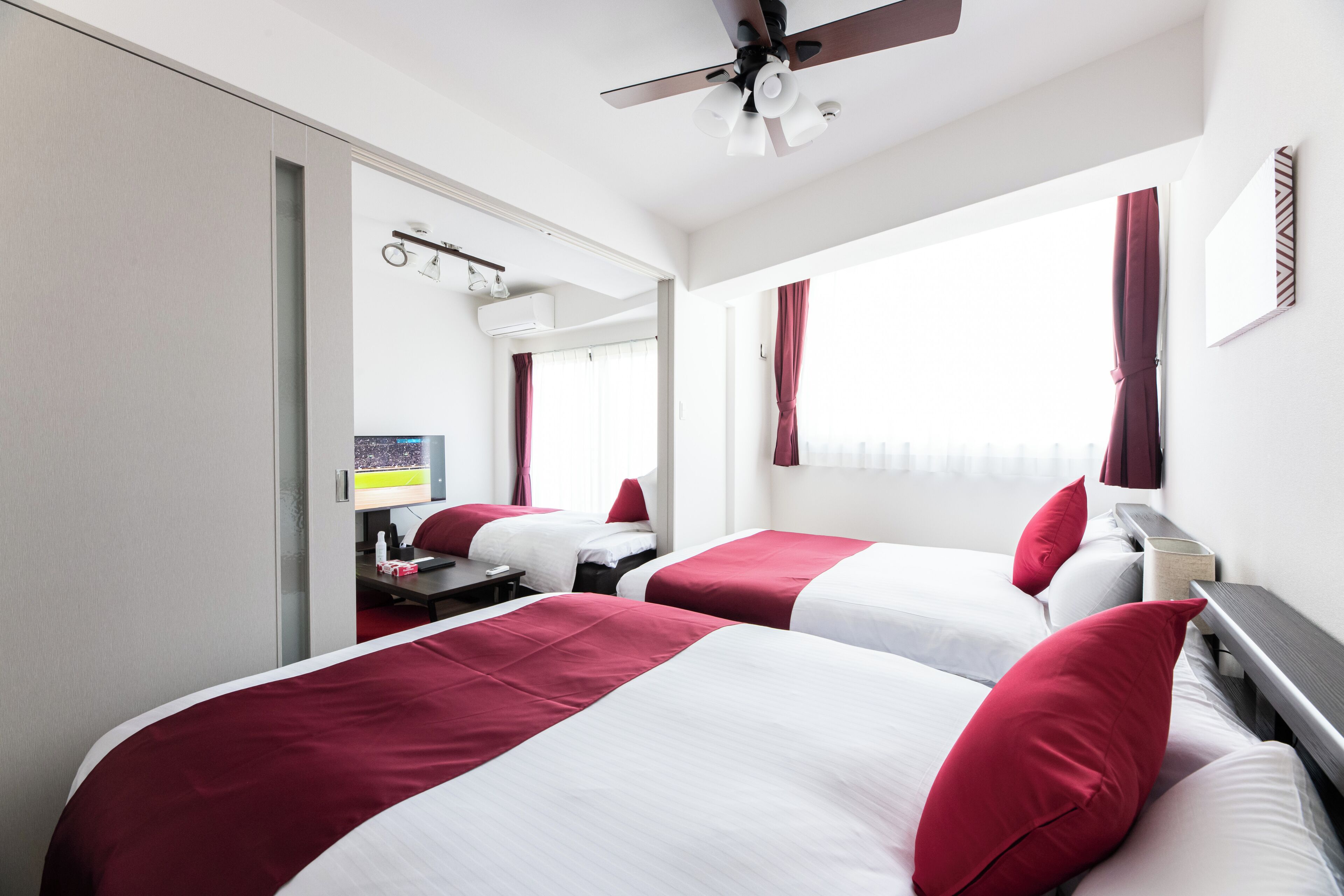 A hotel room with two beds, each with red and white bedding, a ceiling fan, a desk with a computer, and a window with curtains.