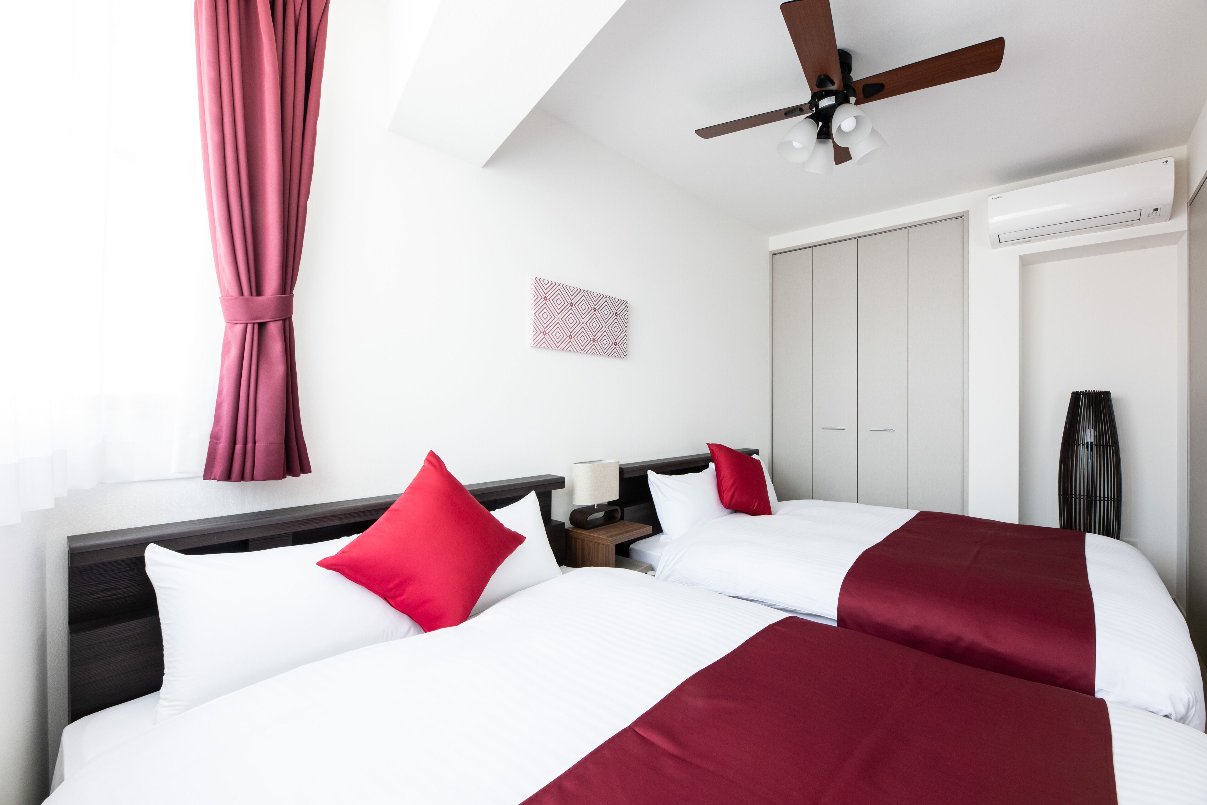 Habitación de hotel con dos camas, ventilador de techo, mesita de noche con lámpara, armario y un elemento decorativo en la pared.