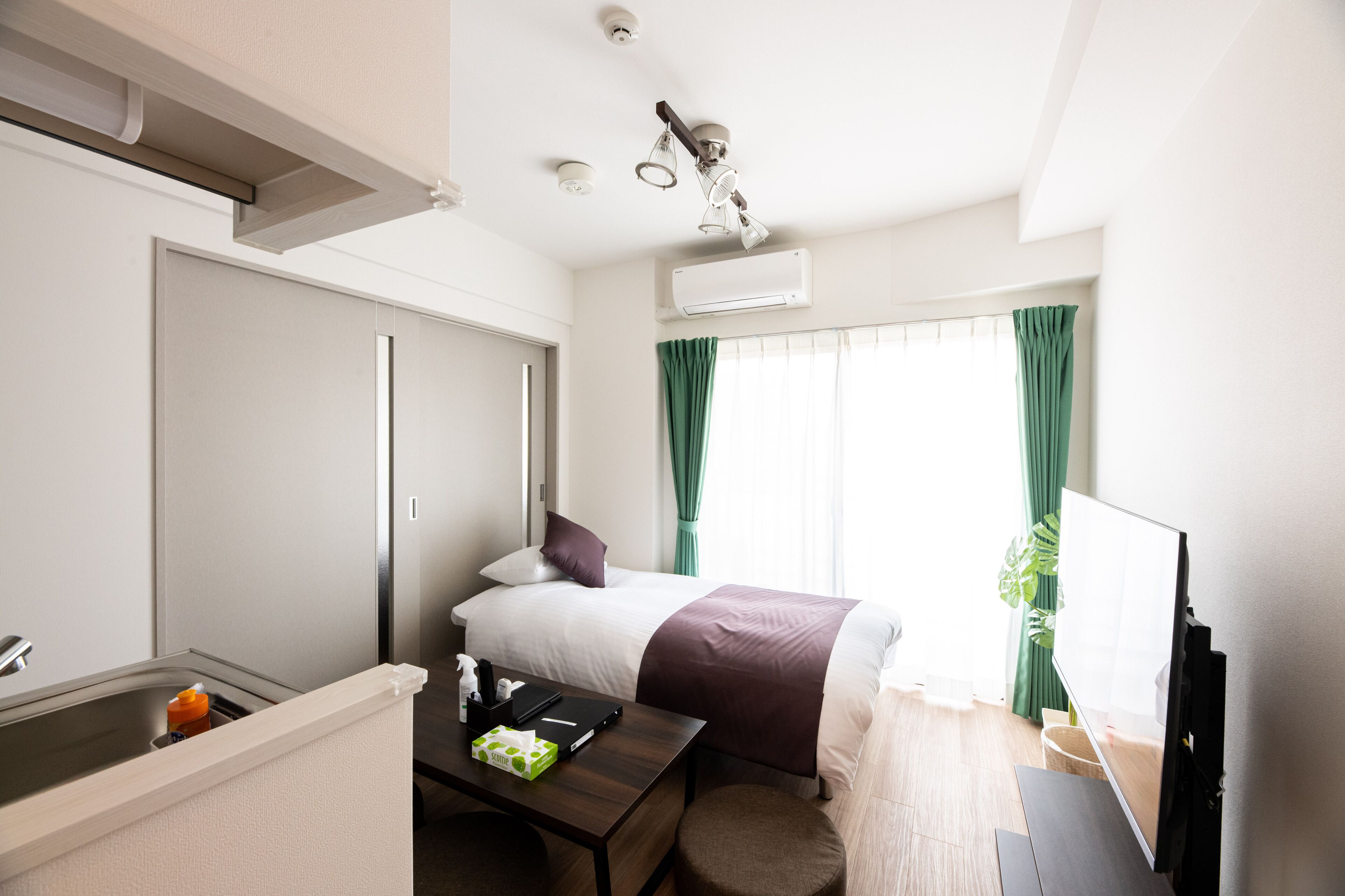 A hotel room with a bed, a desk with a chair, a TV, an air conditioning unit, and a window with curtains.