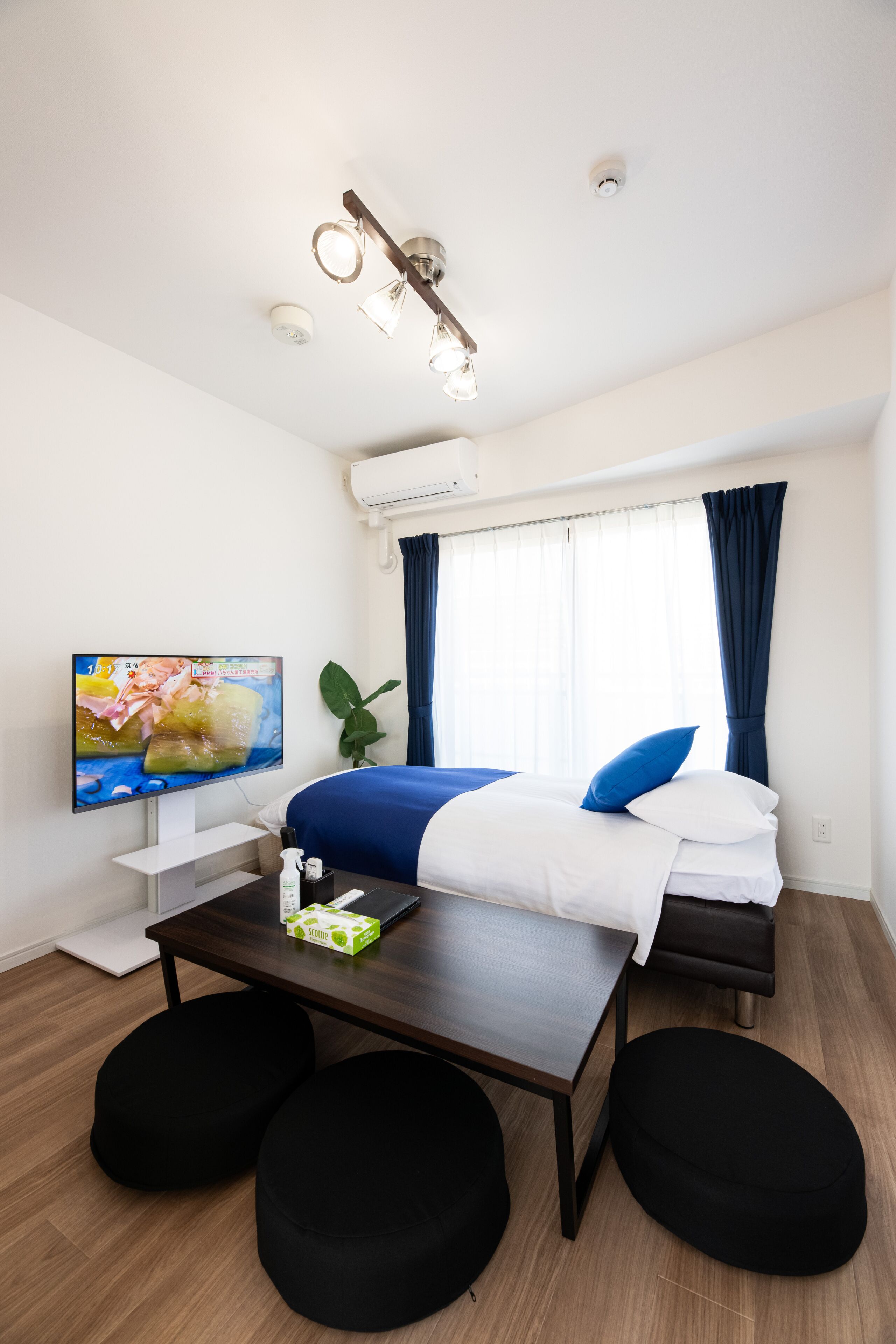 A hotel room with a bed, a TV, a table with a box, and black ottomans.