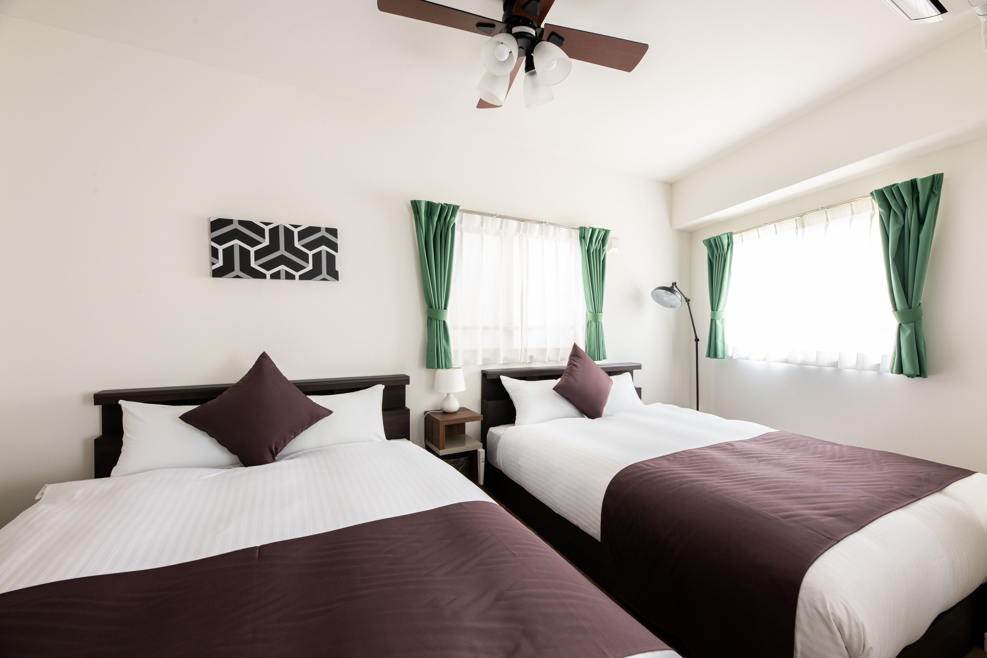 A hotel room with two beds, a ceiling fan, a nightstand with a lamp, and a window with green curtains.