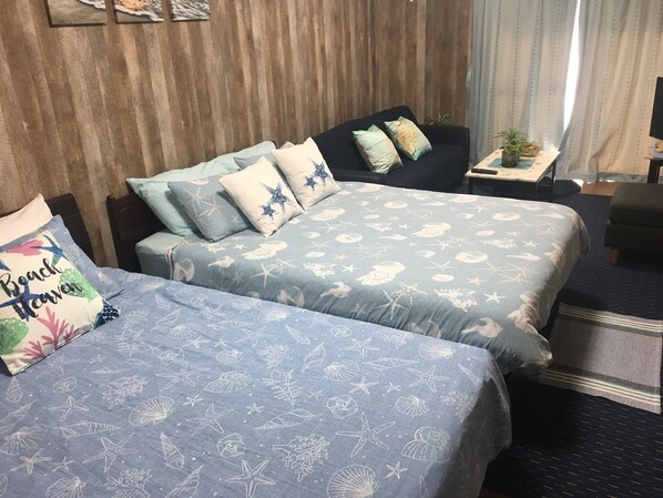 Habitación de hotel con dos camas, cada una con una colcha azul que presenta motivos marinos. Se observa un cabecero de madera, una mesita con una planta y una ventana con cortinas.