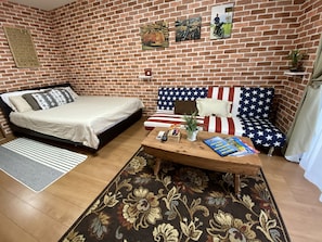 A room with a brick wall, a bed, a sofa with an American flag design, a coffee table with books and a plant, and a small shelf with a clock.