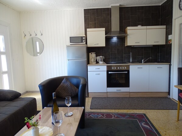 Una cocina compacta con armarios blancos, un microondas, una estufa y un lavamanos. Hay un área de comedor con una mesa, dos copas de vino y una botella.