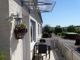 A balcony with a table and chair, a hanging flower pot, and a view of the street and greenery.