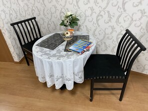Uma mesa redonda com toalha de renda branca, duas cadeiras pretas, um livro, um pequeno vaso com flores e uma cesta com lanches.