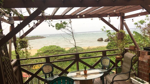A covered patio with a view of the ocean, featuring a brick barbecue, outdoor seating, and hanging plants.