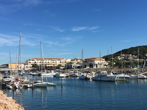 A marina with various boats docked, a building with multiple balconies, and a hilly landscape in the background.