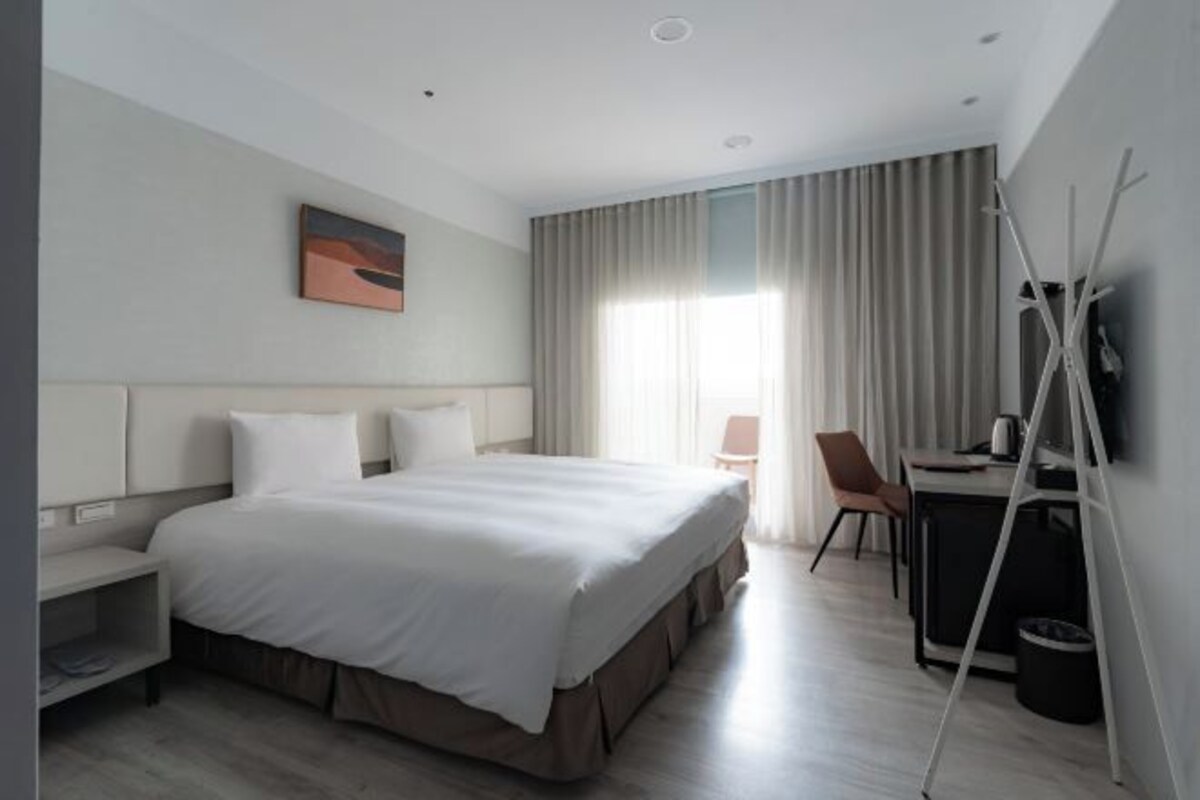 Una habitación de hotel moderna con una cama grande, un escritorio con una silla, televisión y ventana con cortinas.