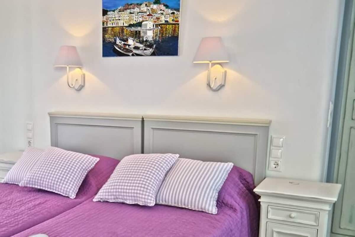 A double bed with a purple bedspread and two pillows, a white nightstand with a lamp, and a wall-mounted picture of a coastal town.