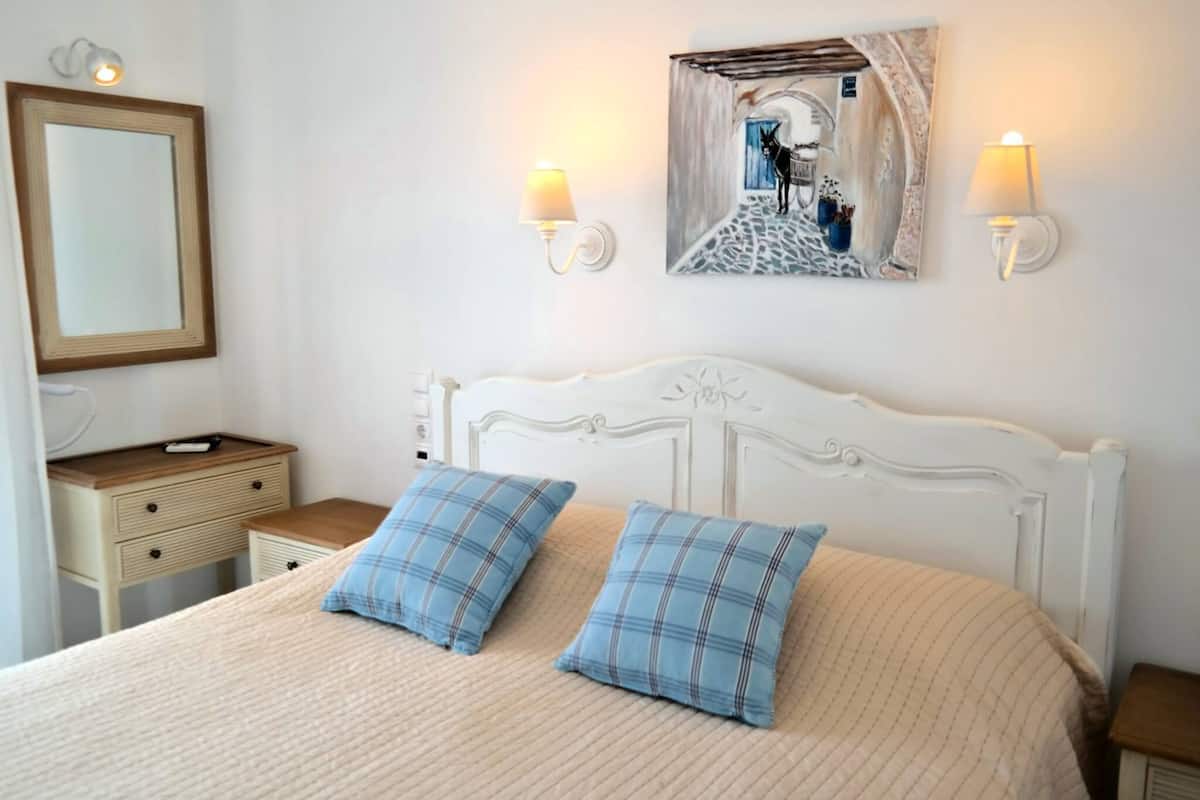 A bedroom with a white headboard, two blue plaid pillows, a beige bedspread, a nightstand with a lamp, a mirror, and a wall art piece.