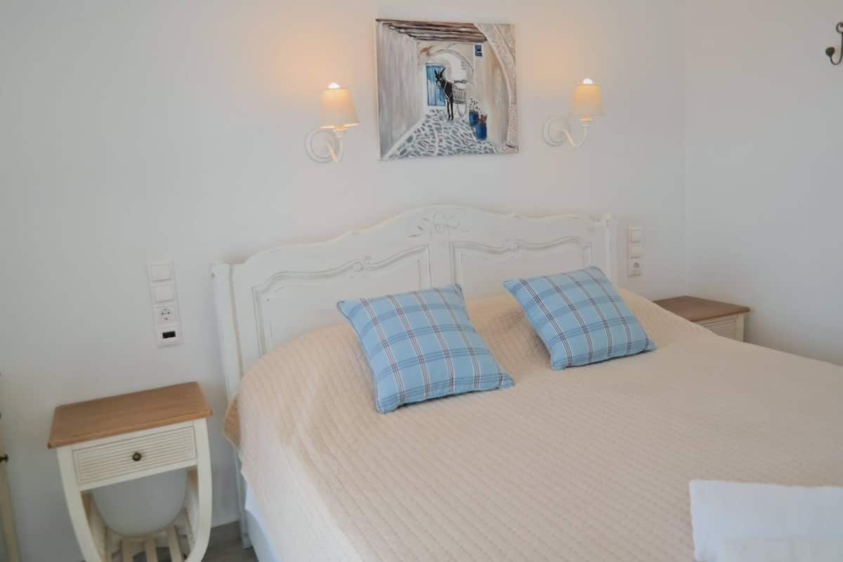 A bed with a white headboard, two blue plaid pillows, and a beige bedspread. There are two bedside tables, one with a wooden top and the other with a white top. A wall-mounted light fixture is visible above the bed.