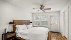 A bedroom with a wooden bed, white bedding, a nightstand with a lamp, a ceiling fan, and a window with blinds.