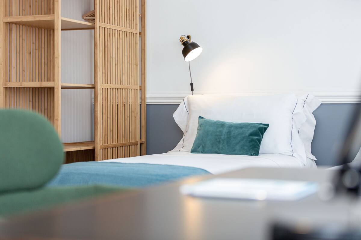 A neatly arranged bedroom with a bed, a bedside lamp, a wooden wardrobe, and a green chair.