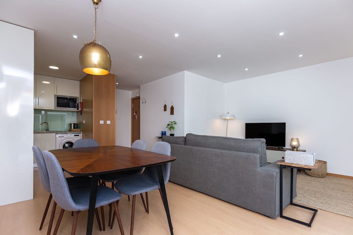 A modern living space with a dining area, sofa, TV, and kitchenette.