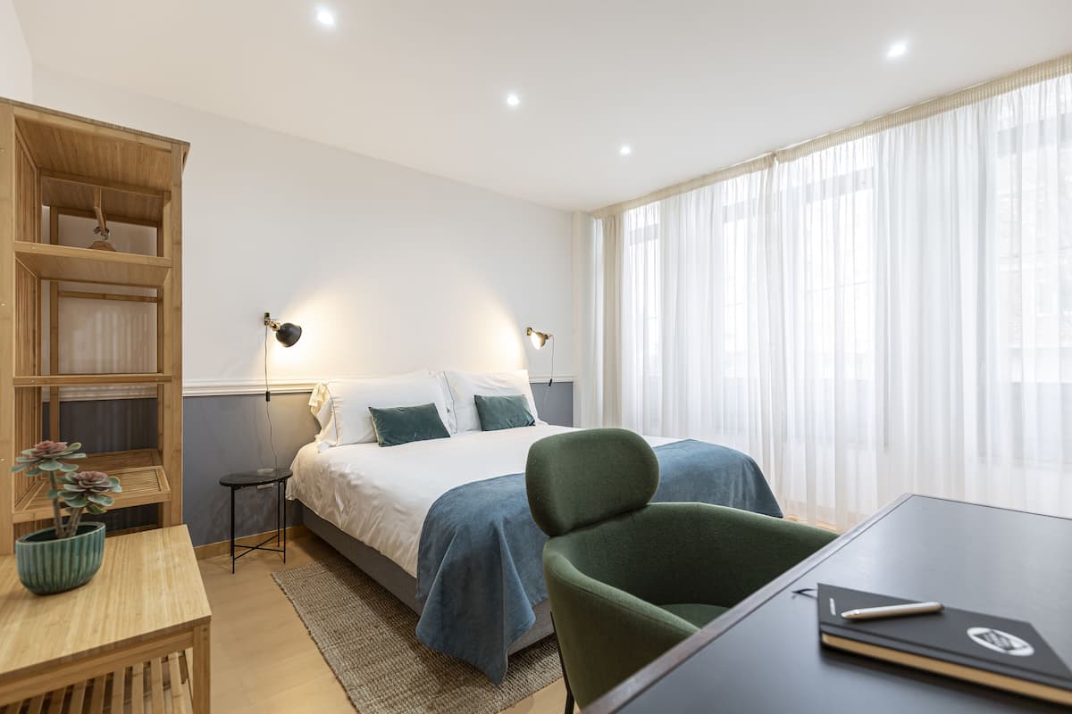A hotel room with a bed, bedside tables, a desk, a chair, a bookshelf, and a window with curtains.