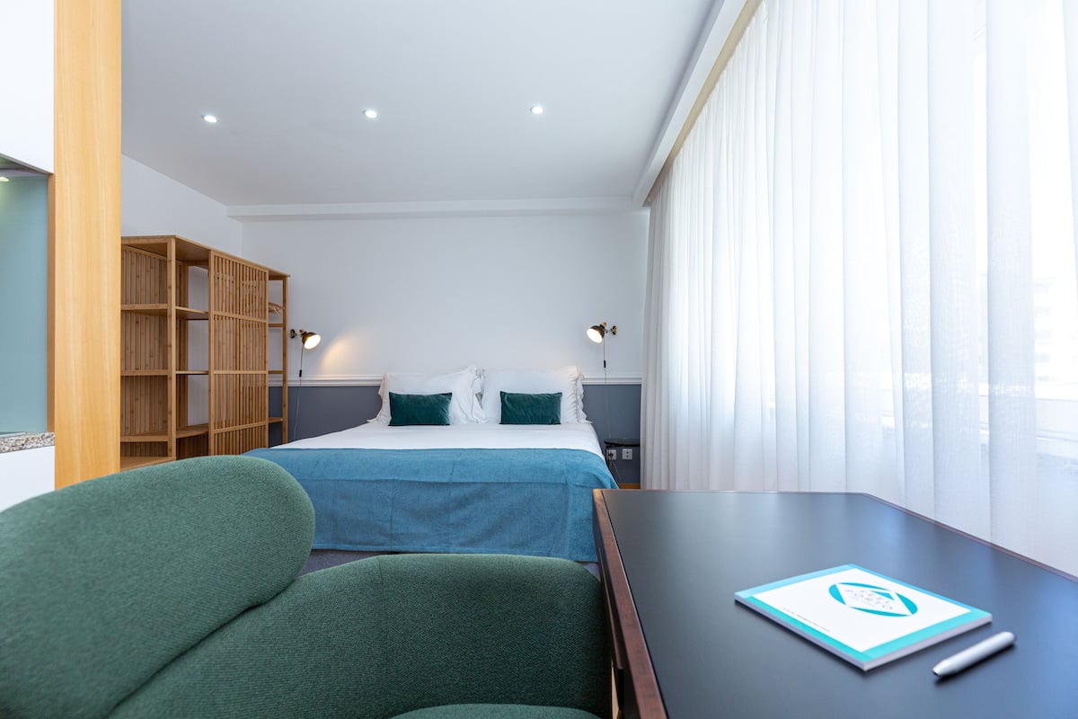 A hotel room with a bed, a desk, a chair, a bookshelf, and a window with curtains.