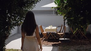 Mulher de vestido branco caminhando em direção a uma mesa e cadeiras ao ar livre.