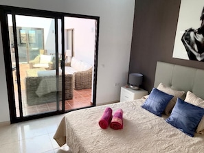 A bedroom with a bed, pillows, a nightstand, a lamp, and a view through a sliding glass door to an outdoor seating area.
