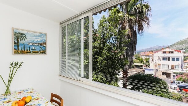 Un balcon avec une table dressée pour le dîner, un tableau représentant un paysage côtier et une vue sur une ville avec des bâtiments et une colline au loin.