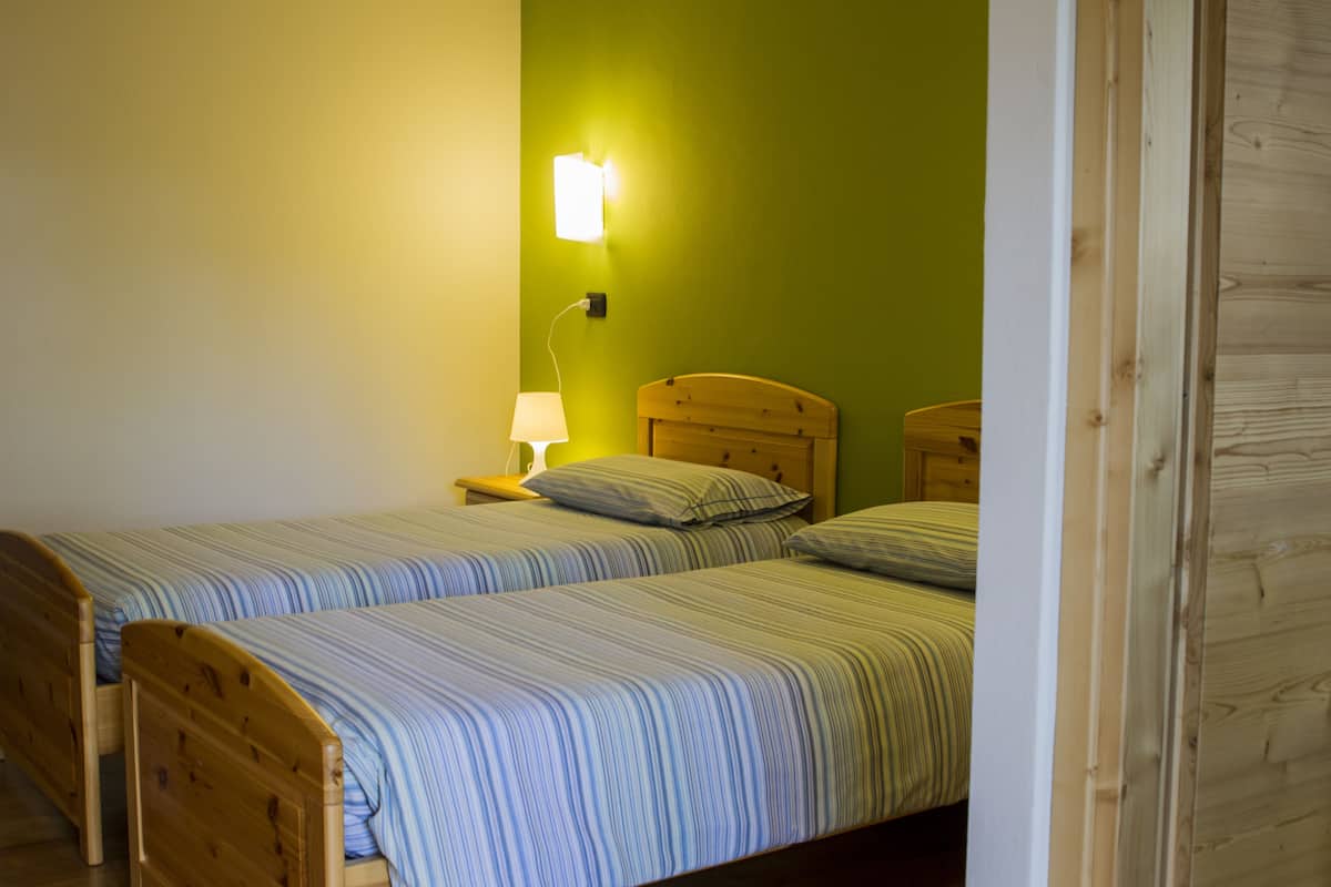 A room with two single beds, each with a wooden headboard and striped bedding. A nightstand with a lamp is visible on the left side.