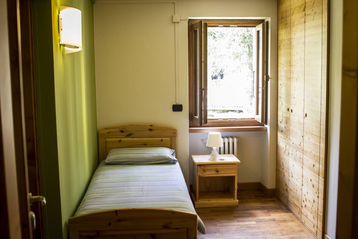 A small, single-bed room with a wooden headboard, a bedside table with a lamp, a window with a view of greenery, and a radiator under the window.