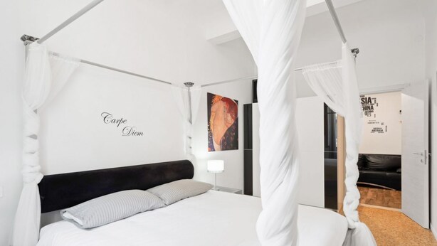 A modern bedroom with a four-poster bed, a nightstand, a wall-mounted lamp, and a framed artwork. - GuestHost - Located in the heart of the city, near the places of greatest cultural interest and a few steps from the Santa Maria Novella station, it is the ideal place for tourist or business stays.The area of ​​Borgo Ognissanti reflects the ancient  (Florence)