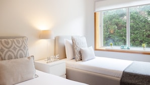 Quarto com duas camas, uma mesa de cabeceira com abajur e janela com vista para o verde.