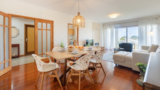 Uma sala de estar moderna com uma mesa de jantar de madeira, cadeiras brancas, um sofá e uma grande janela com cortinas.
