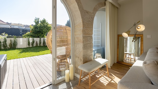 A modern living room with a wooden floor, a white sofa, a bench, and a wicker chair. There is a large window with a view of a garden and houses.