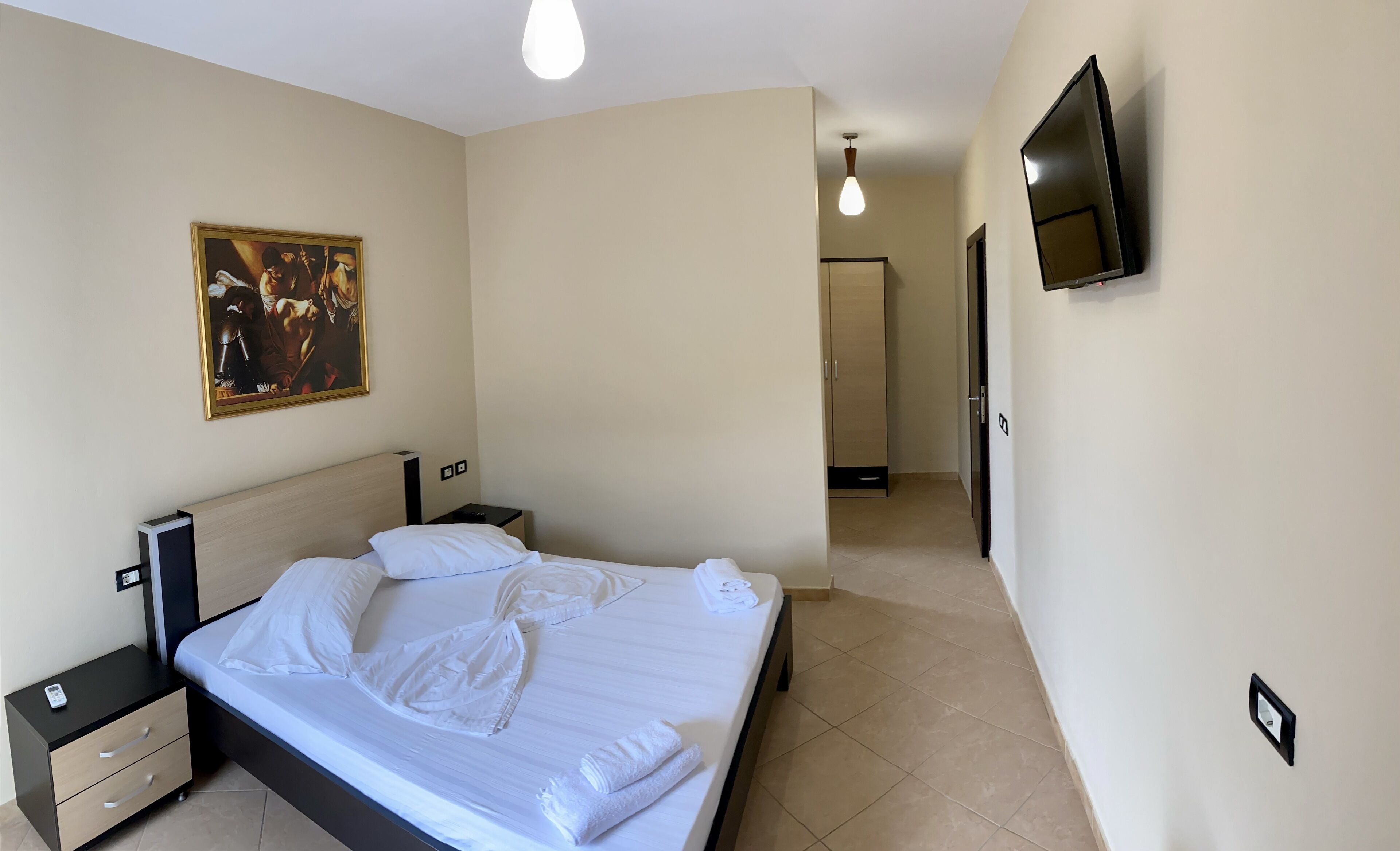 A hotel room with a bed, a nightstand, a painting, a TV, and a hallway.