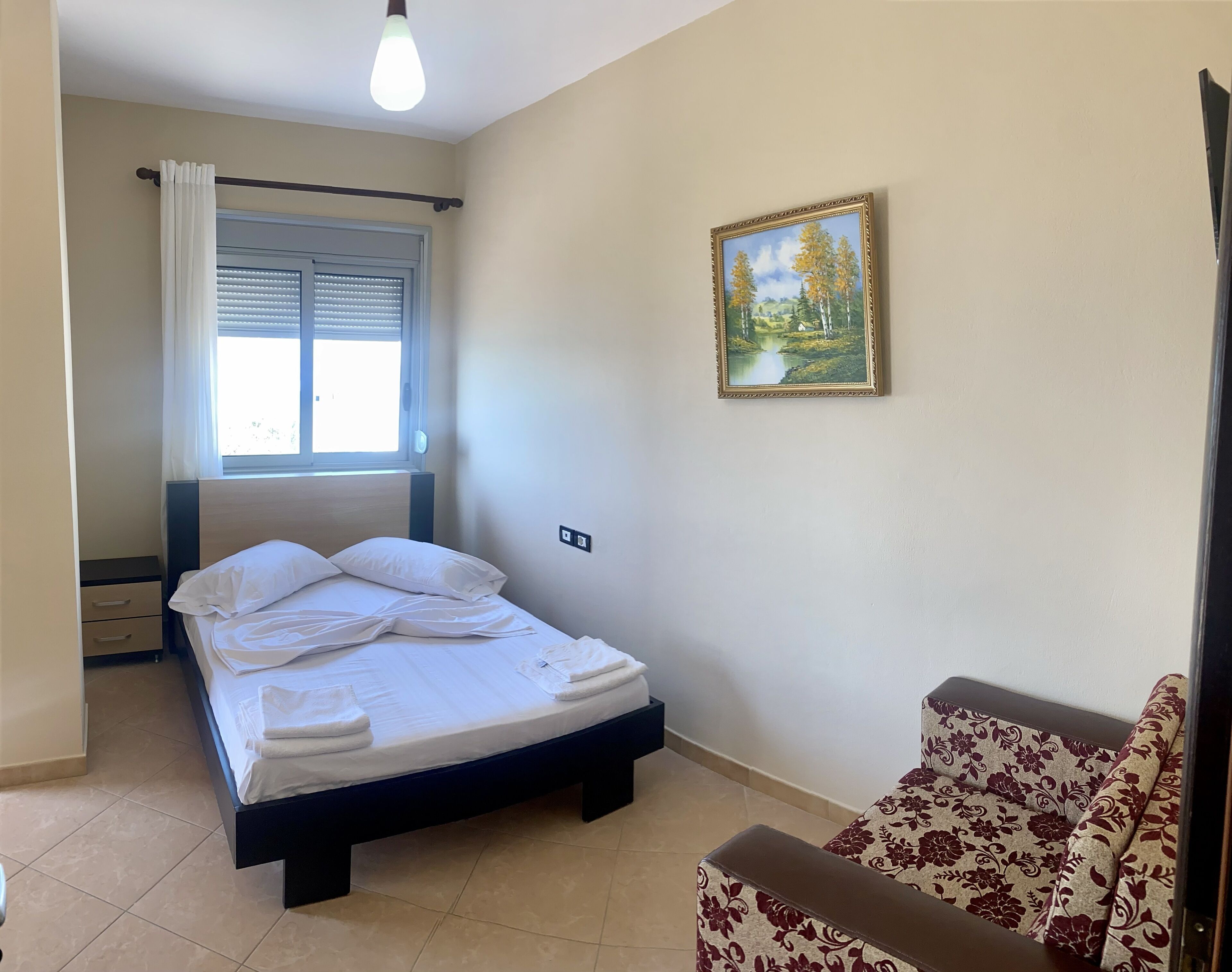 A bedroom with a bed, a window with blinds, a painting on the wall, and a sofa.