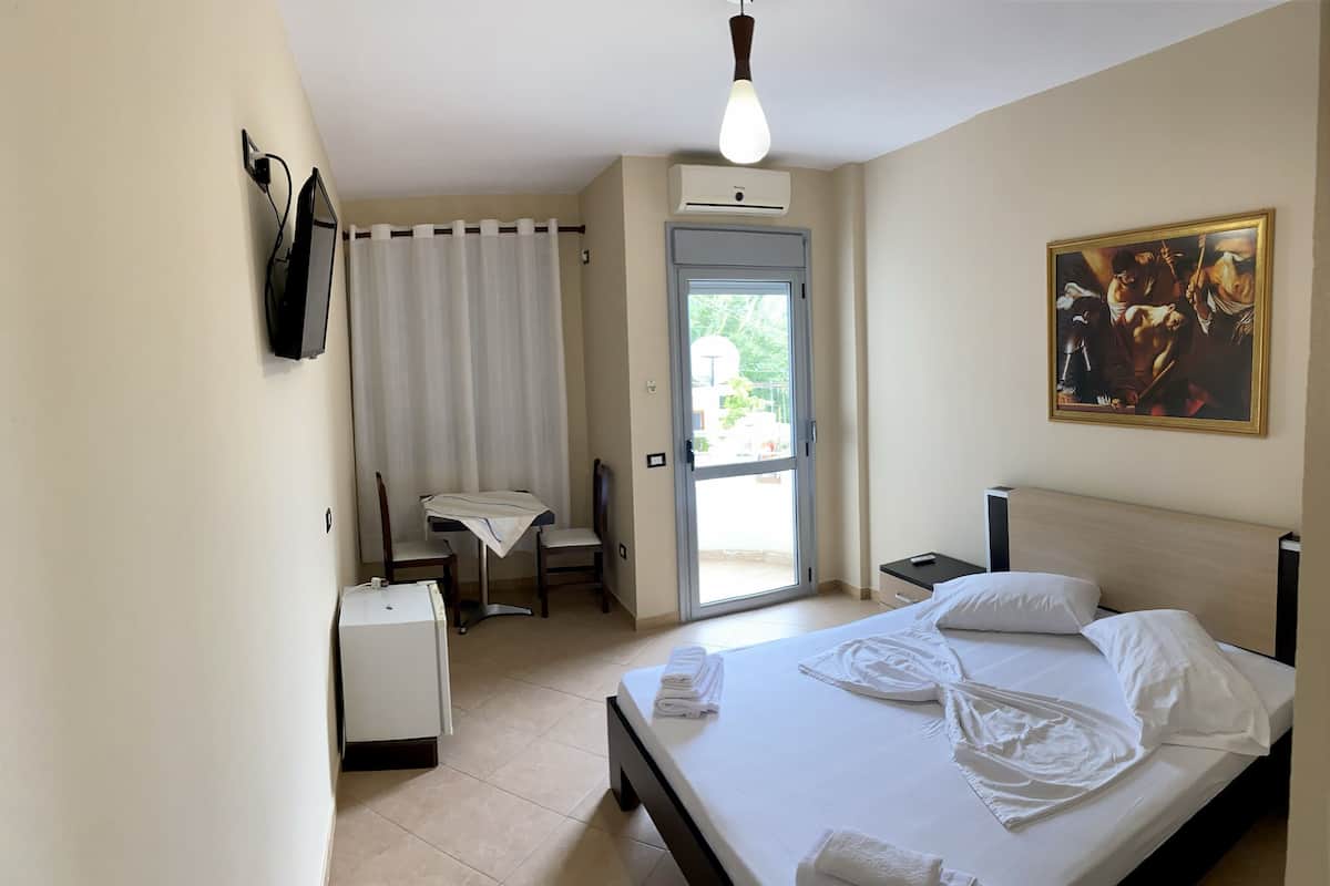 Quarto de hotel com uma cama, uma escrivaninha, uma cadeira, uma mesinha, televisão na parede e um quadro.