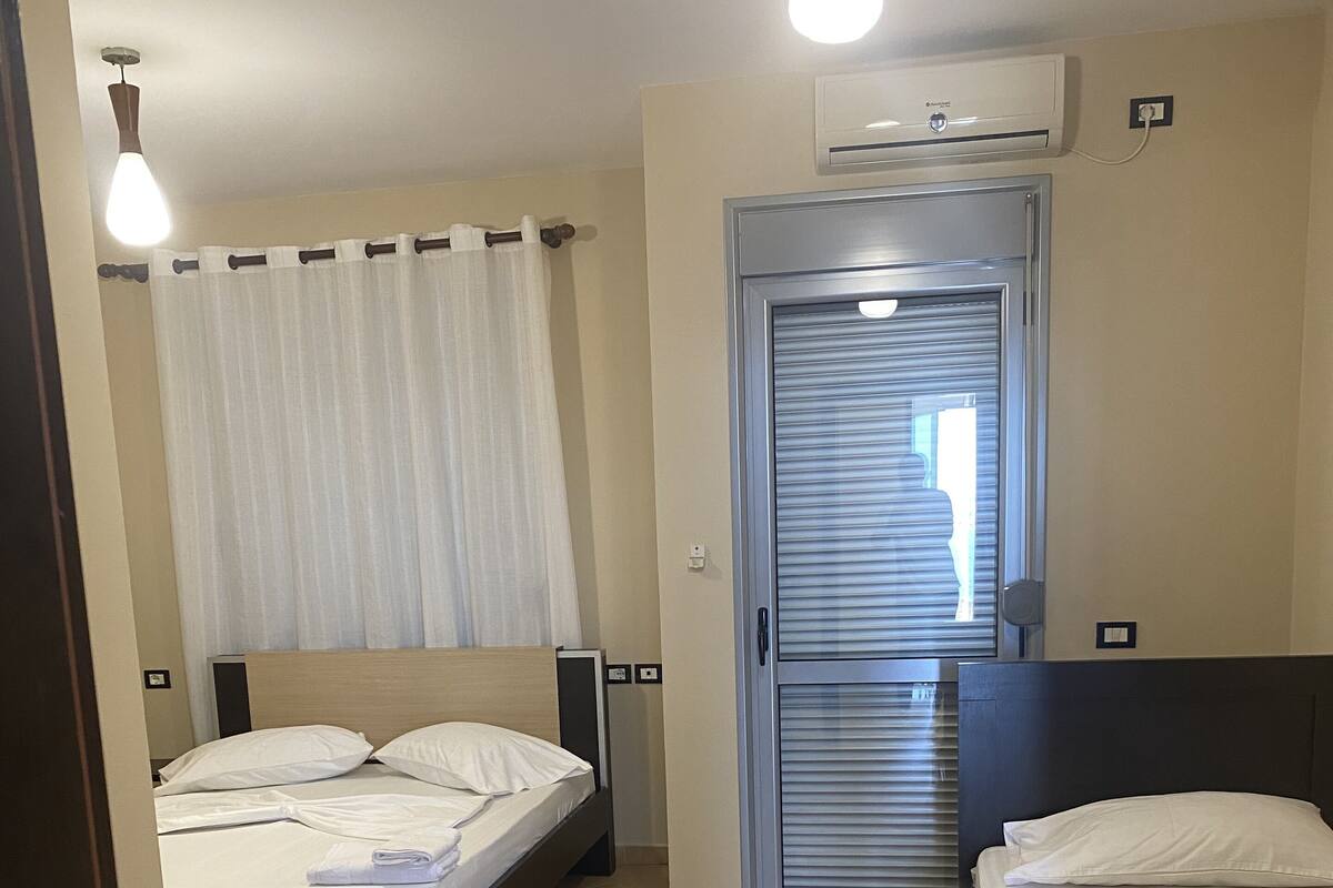 Quarto de hotel com duas camas, ventoinha no teto, aparelho de ar condicionado e porta deslizante.