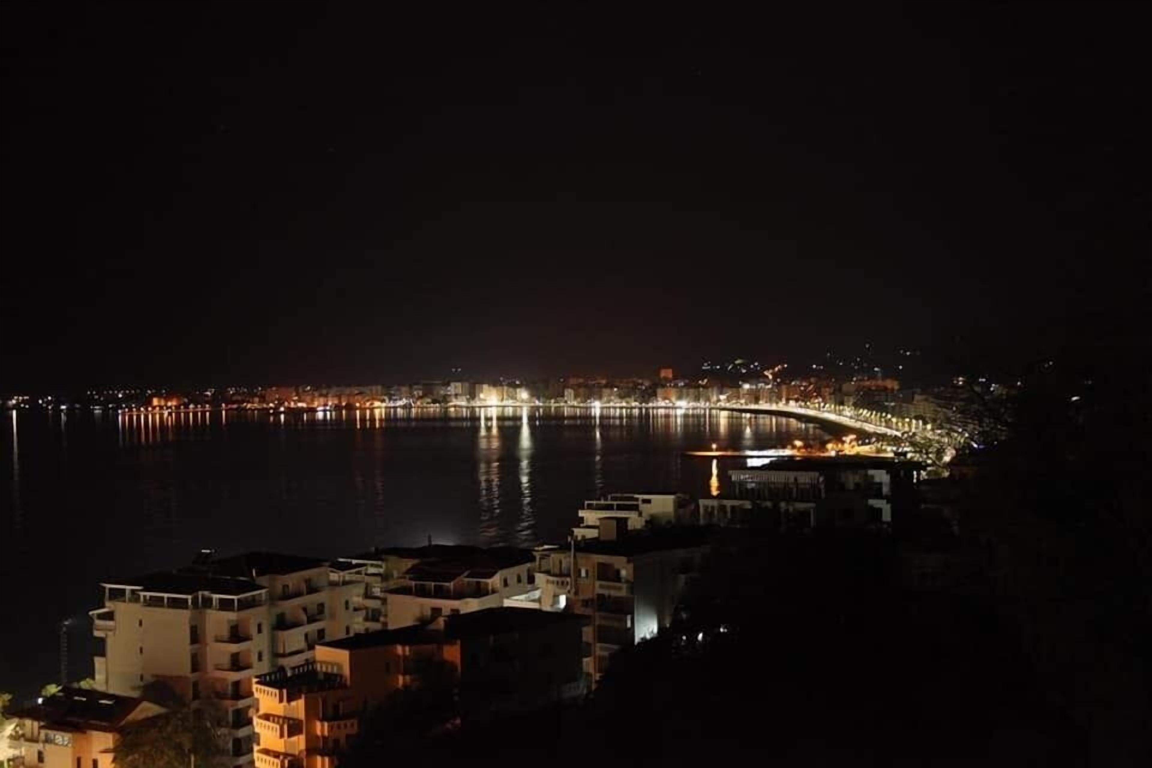 A nighttime view of a coastal city with a brightly lit waterfront promenade and a bridge.