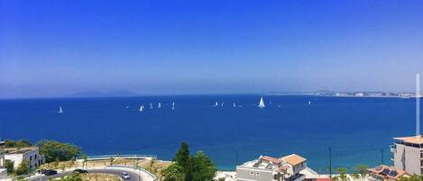 A coastal view with buildings, a road, and sailboats on the sea.